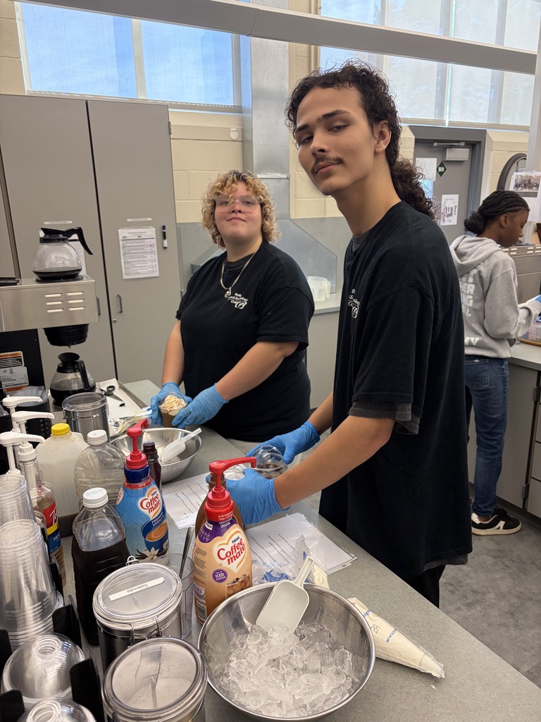Two people in black shirts and gloves prepare food in a kitchen with bottles and containers.
