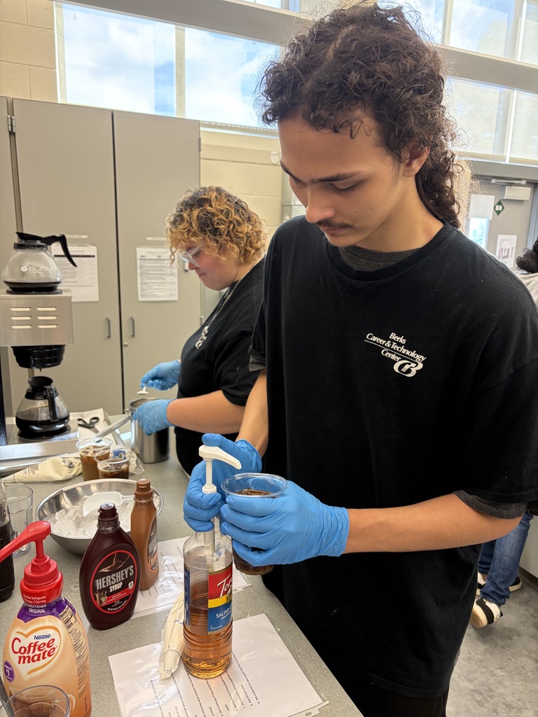Two people prepare food in a lab, one holding a bottle with a pump. One wears glasses and a black shirt, the other blue gloves.