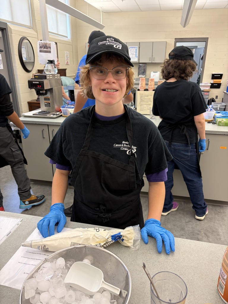 A young person in a black shirt, cap, and blue gloves works at a counter, surrounded by others.
