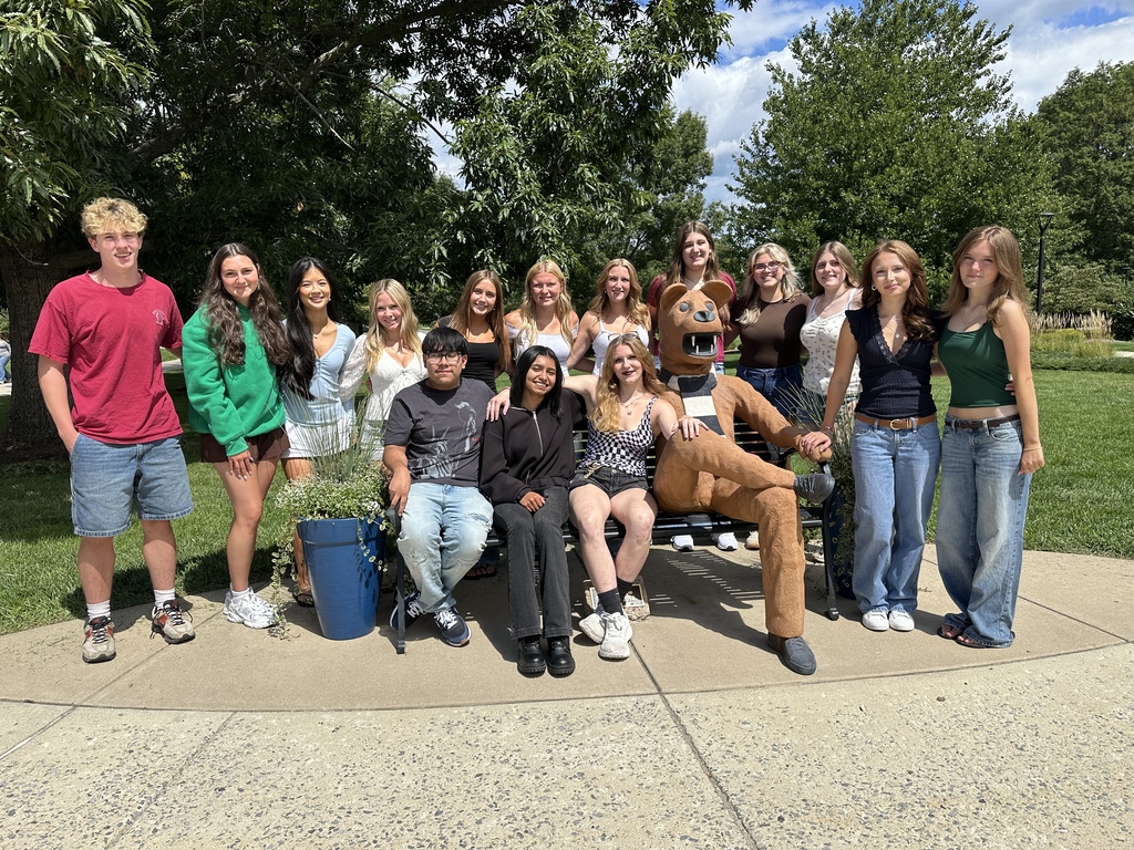 A group of students stands on a sidewalk, smiling, with one in a mascot costume. Trees and grass are in the background.