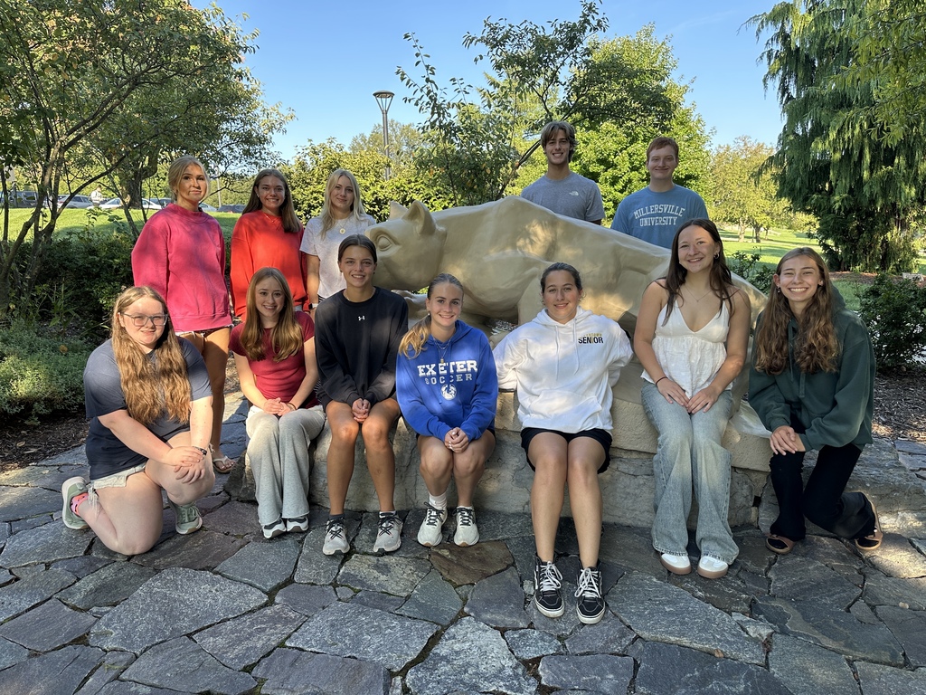 Group of people smiling, some sitting on a stone bench, posing for a photo. Trees and bushes in background.