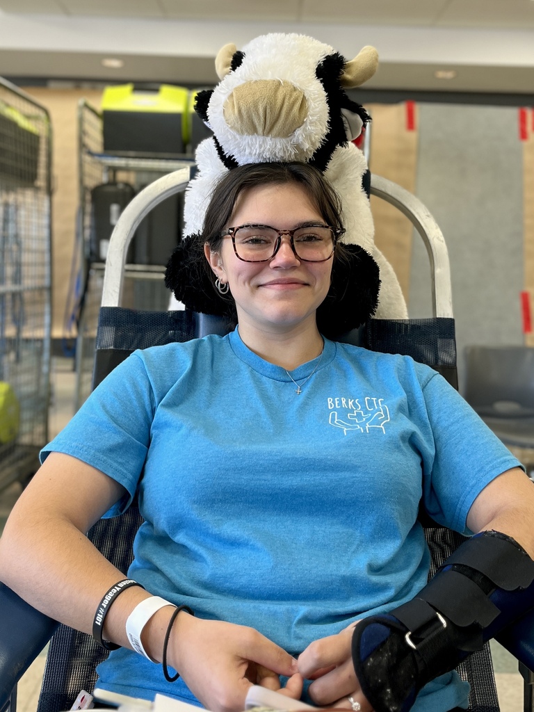 A woman with a bandaged arm sits in a chair wearing a blue shirt. A cow plush rests on her head.