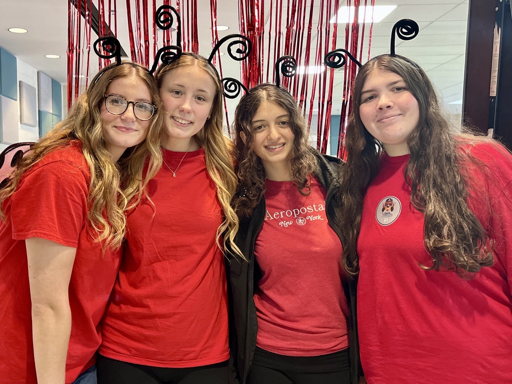 Five women wearing red t-shirts stand together with smiles, against a backdrop of red streamers.