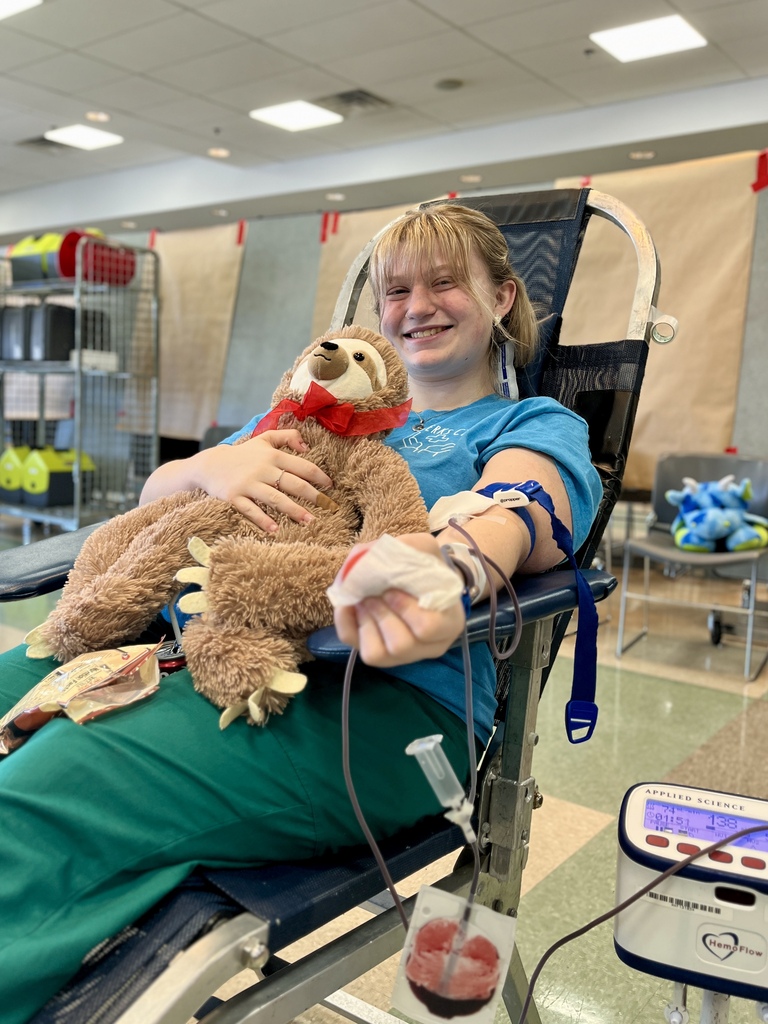 A young woman in green scrubs, sitting in a chair, holds a stuffed animal while receiving treatment.