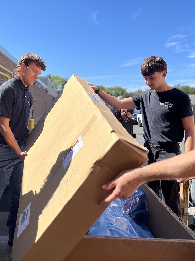 Two young men in black shirts lift a large box while another person assists. Blue sky in background.