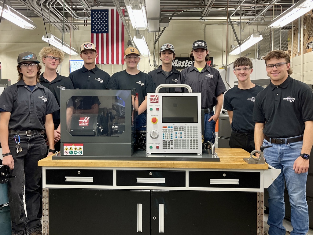 Nine individuals, in uniform, stand near a large machine in a workshop. One person is touching the machine.
