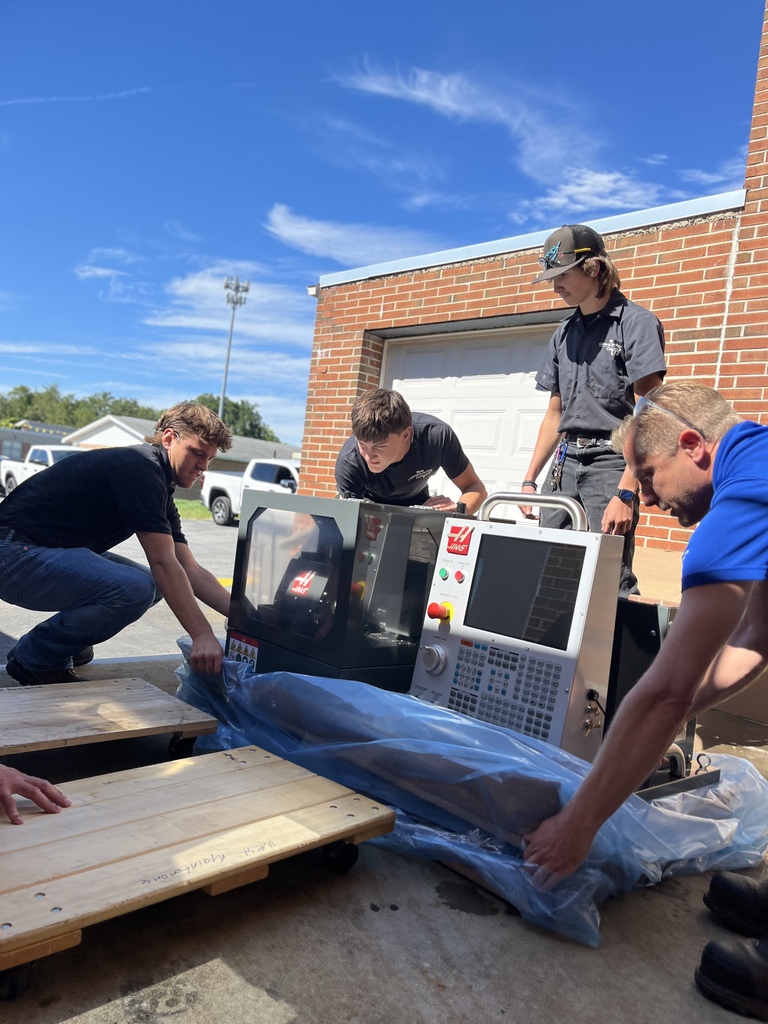 Five people are unloading a machine from a cart. They are working on a paved area.