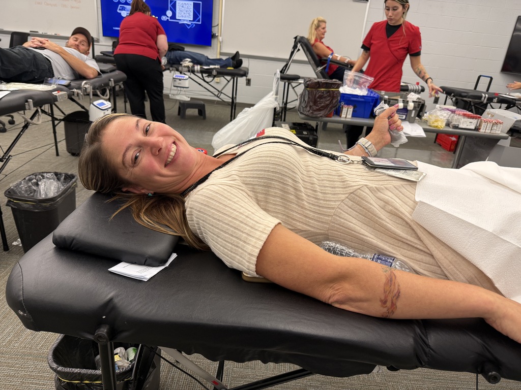 A smiling woman lying on a bed, likely at a blood donation event, with others in red shirts in background.