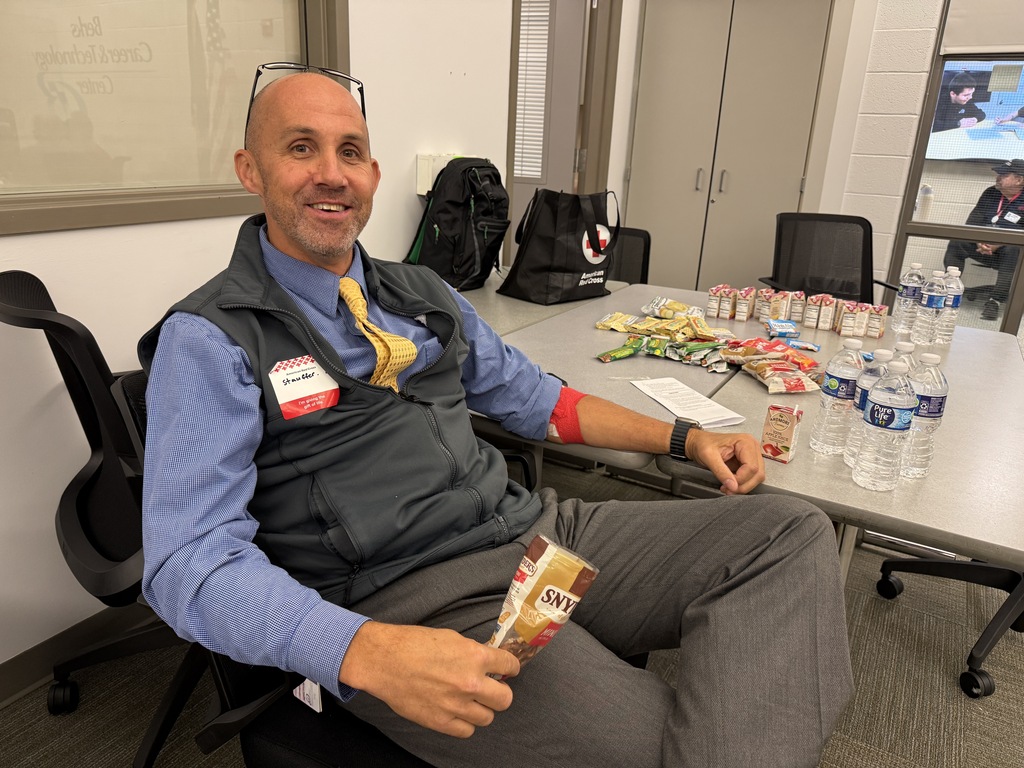 Man sitting in chair at desk holding snack. Many snacks, water bottles, and items on table.