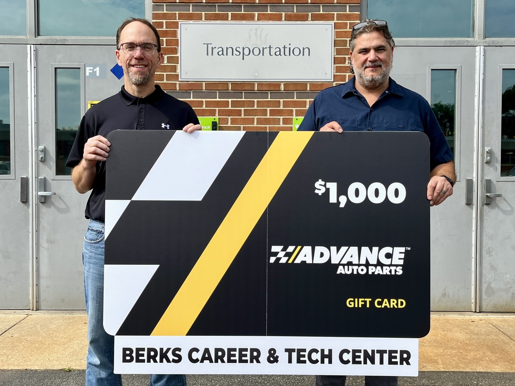 Two men hold a large sign with a $1,000 gift card from Advance Auto Parts, in front of a brick building.