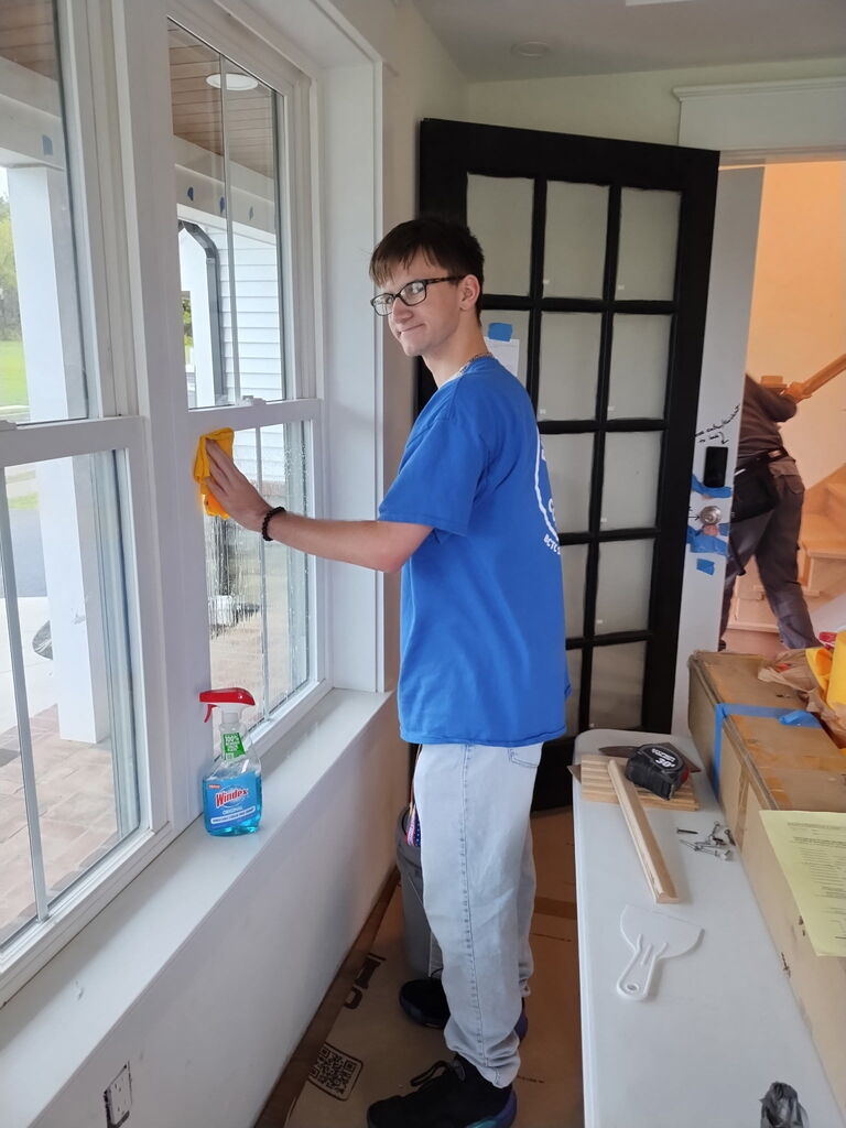 A man in a blue shirt and glasses cleans a window with a yellow cloth. Behind him, a door leads to another room.