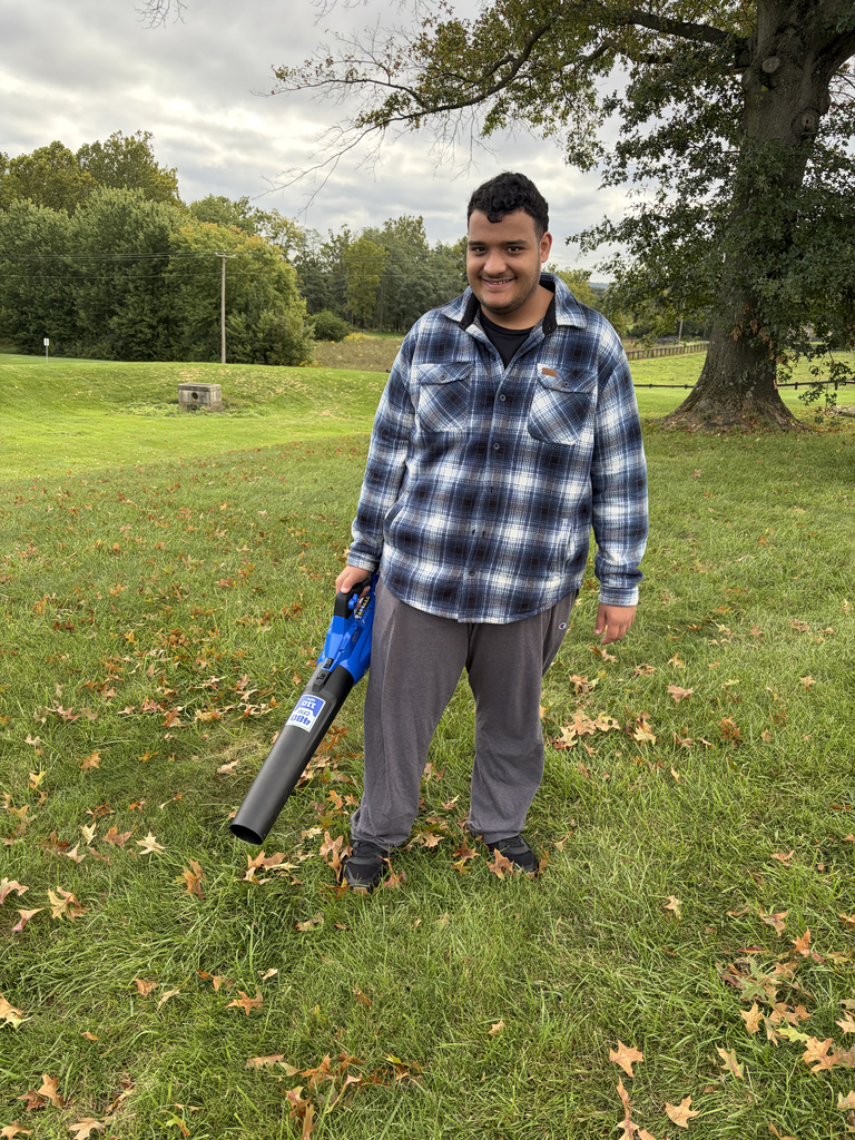 Person in a plaid shirt and gray pants holding a leaf blower in a grassy field.