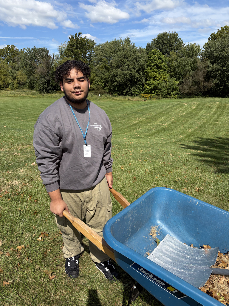 Person holding a blue wheelbarrow with a flat bottom filled with debris in a grassy field.