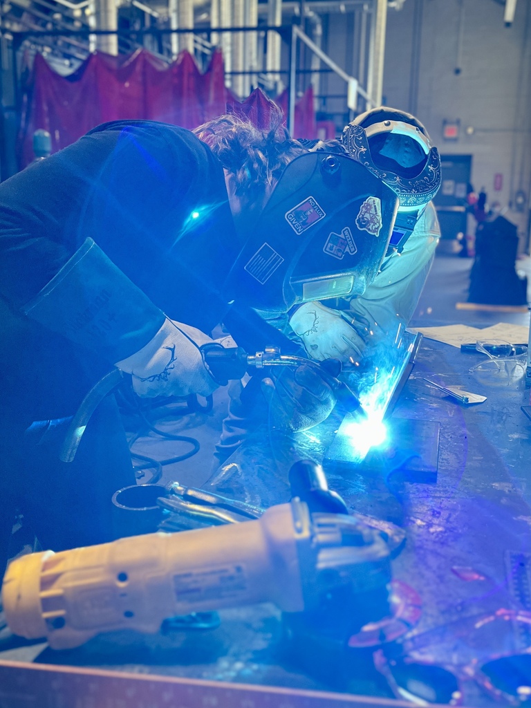 Two people in safety gear weld together metal in a workshop with blue and red lighting.