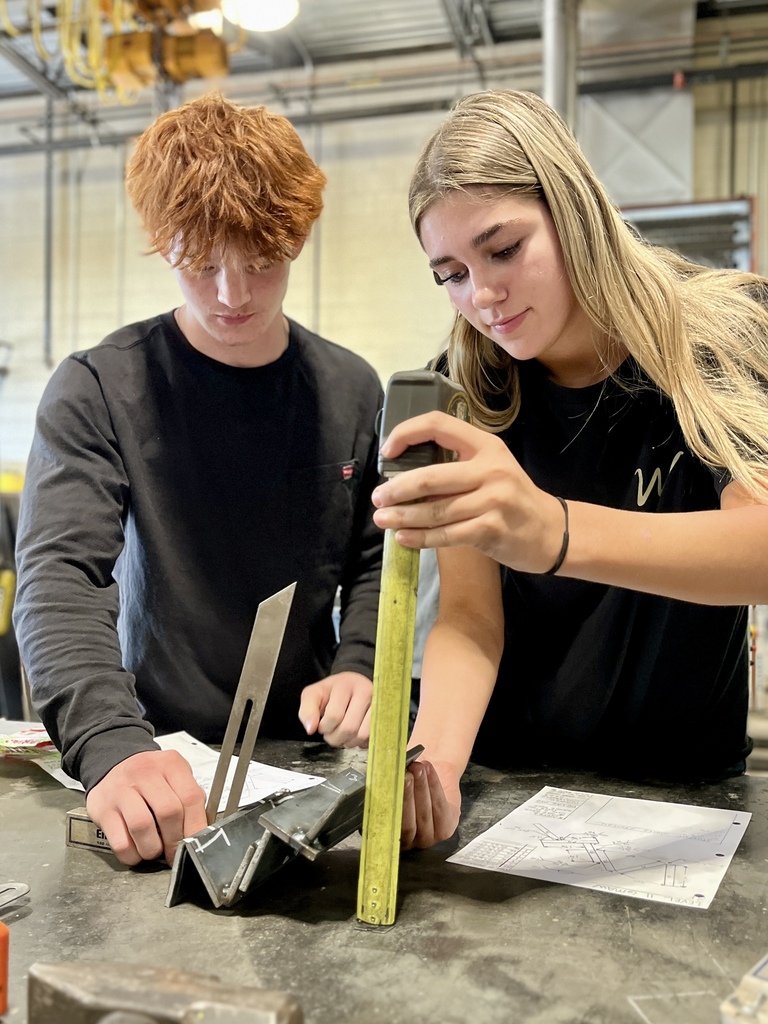 Two people work at a table, examining a metal part. One holds a measuring tape, and the other holds a square.