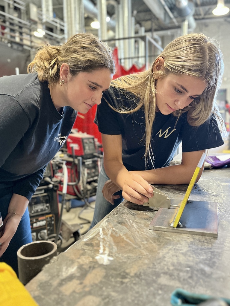 Two women examine a metal sheet on a table in an industrial setting. One holds a piece of paper.