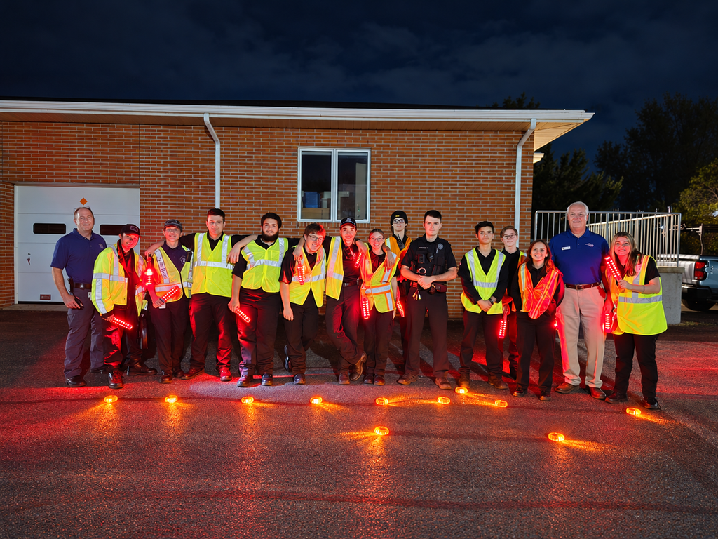 A group in high visibility gear poses in front of a brick building at night, with red lights on the ground.