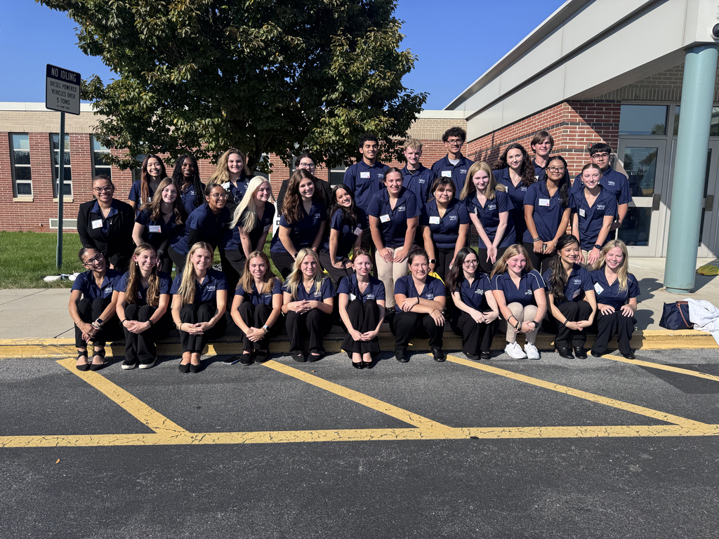 Group of students in matching blue shirts, sitting and standing in rows in a parking lot.