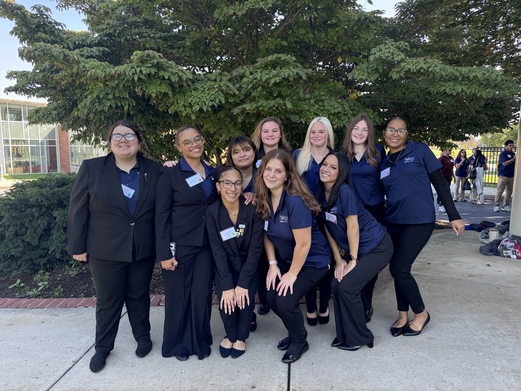 Eleven people in blue shirts and black pants pose for a photo in front of a tree.