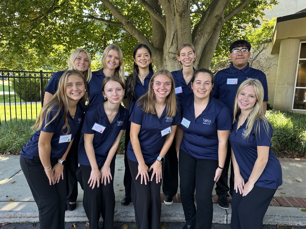 A group of students dressed in blue shirts and black pants pose for a picture outside.