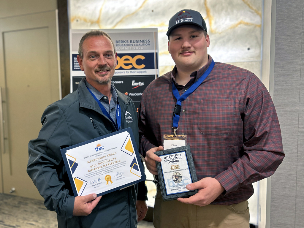 Two men standing, one wearing a cap, both holding awards and wearing lanyards, in a room with a marble wall.