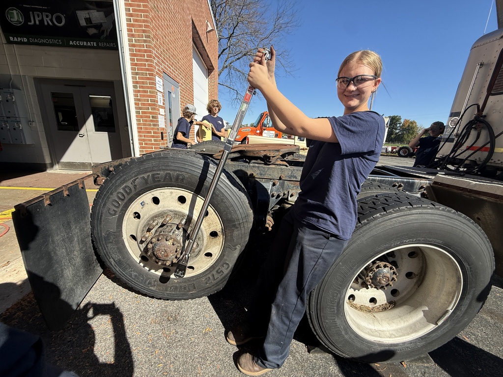 A student holds a wrench next to a large tire on the back of a truck.