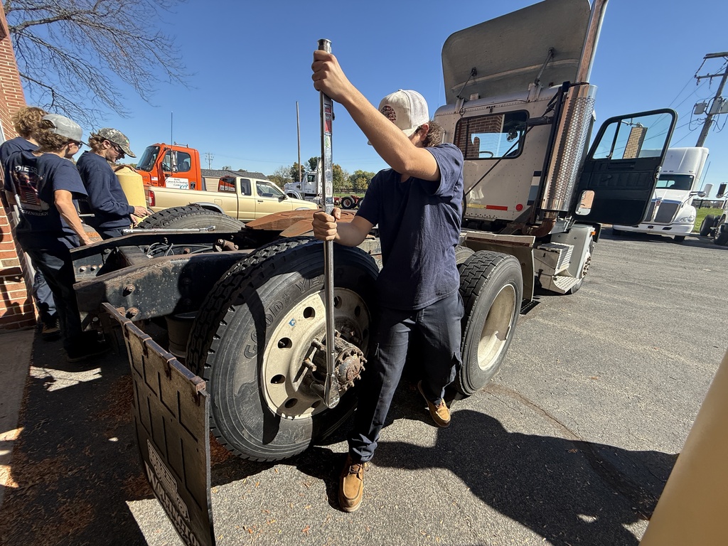 Students examine a semi-truck's tire and undercarriage with a tool while standing in a parking lot.
