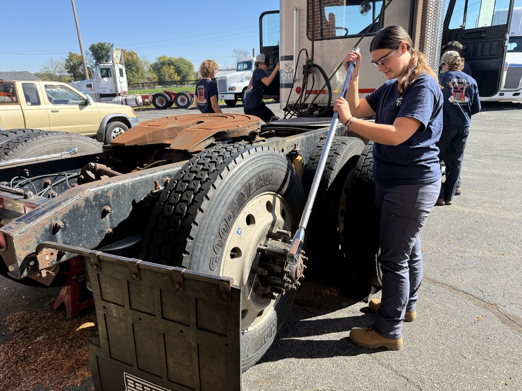Woman working on the rear of a large vehicle with a large metal tool. Three people in background.