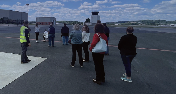 A group of individuals look towards an airplane strip as an instructor in a safety vest provides an educational tour.
