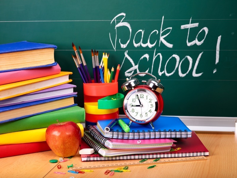 Stack of colorful books, notebooks, and school supplies including pencils, pens, scissors, and paperclips on a desk in front of a chalkboard with the words 'Back to School!' written in chalk. An apple and a red alarm clock are also placed on the desk.