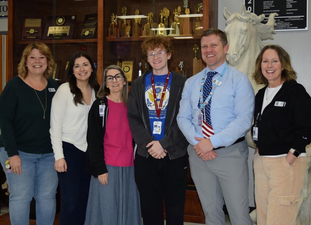 Six people stand together smiling inside Bethany High School in front of a wooden trophy display case filled with plaques, medals, and academic and activity awards. Bethany High School senior Luke Gillespie stands in the center wearing glasses, a blue Bethany shirt, and a school lanyard with ID, surrounded by school staff and administrators on both sides. A large white horse statue, representing the Bethany Bronchos, is visible behind the group on the right. The group is posed for a recognition photo celebrating Luke being named a National Merit Finalist.