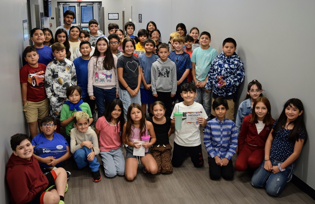 A large group of multilingual learner students gather in a school hallway for a group photo. The students are of various ages, with some holding certificates and cups from their graduation celebration. They are smiling and standing close together, showing excitement and pride.