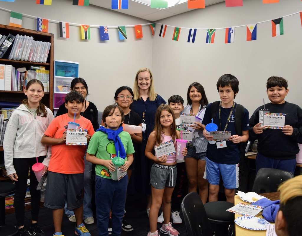 A group of multilingual learner students stand together in a classroom with a teacher, smiling and holding colorful certificates and cups. International flags hang across the top of the room. The students look proud of their accomplishments, and the atmosphere is celebratory.