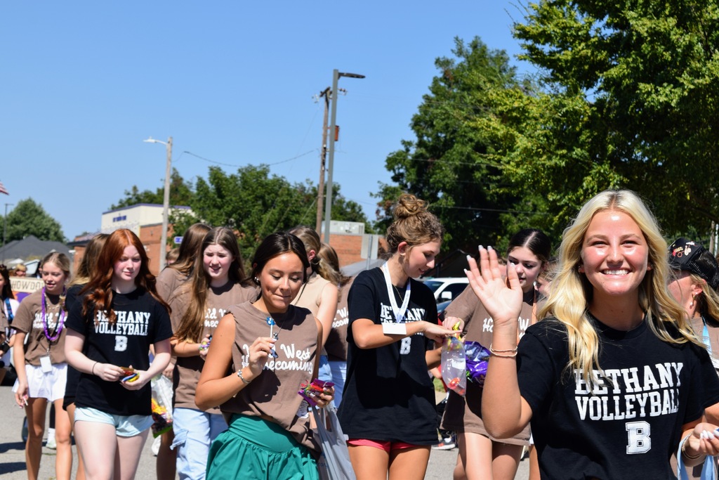 A group of high school volleyball players walk in the parade, smiling and passing out candy. The girl in the foreground waves with a big smile while wearing a black "Bethany Volleyball" shirt. Others in brown "Wild West Homecoming" shirts and black team shirts walk behind her.