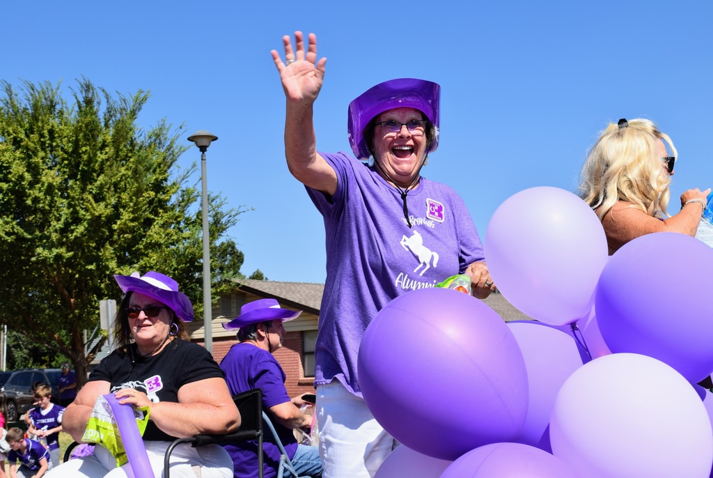 A woman wearing a purple cowboy hat and a purple Bronchos Alumni t-shirt waves energetically from a parade float decorated with purple balloons. She smiles widely, while other women in purple hats and shirts sit beside her.