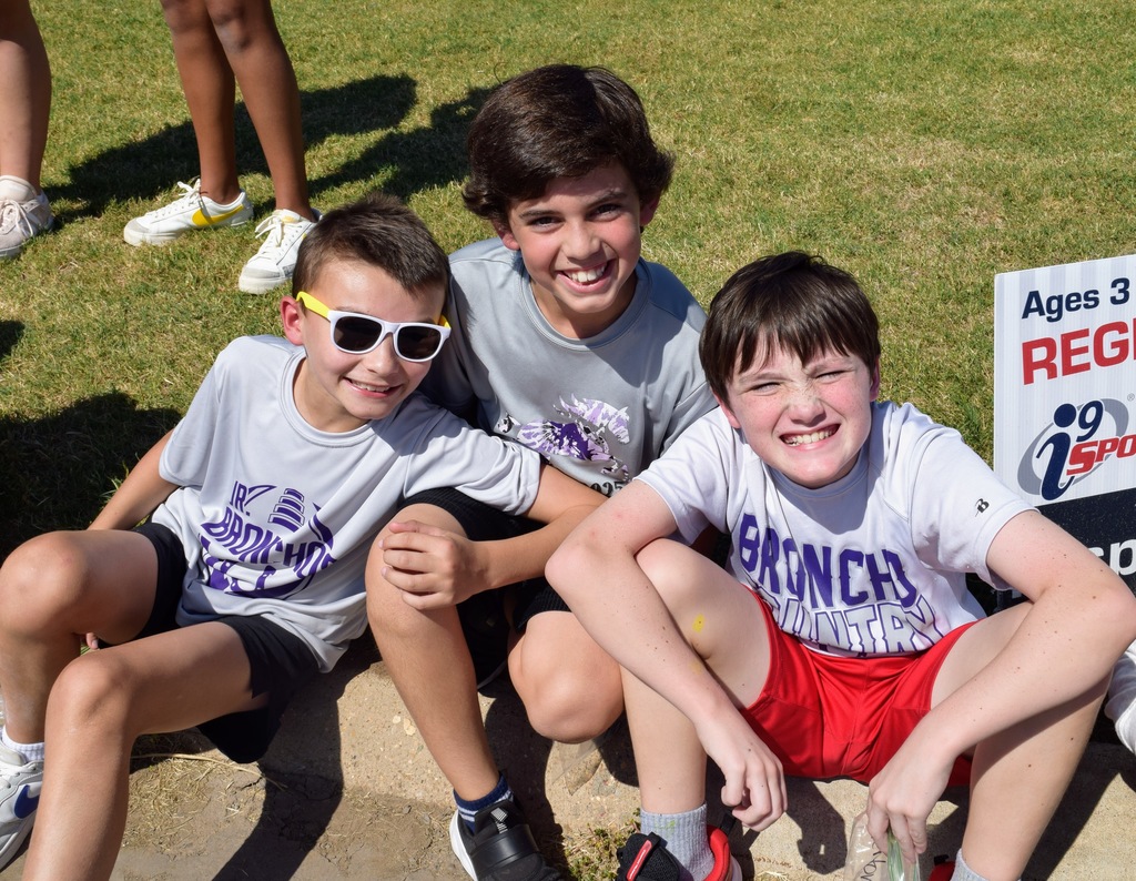 Three young boys sit together on the curb, smiling for the camera. They wear Bethany Bronchos t-shirts, with one boy in sunglasses, another in a gray shirt, and the third in a white "Broncho Country" shirt with red shorts.