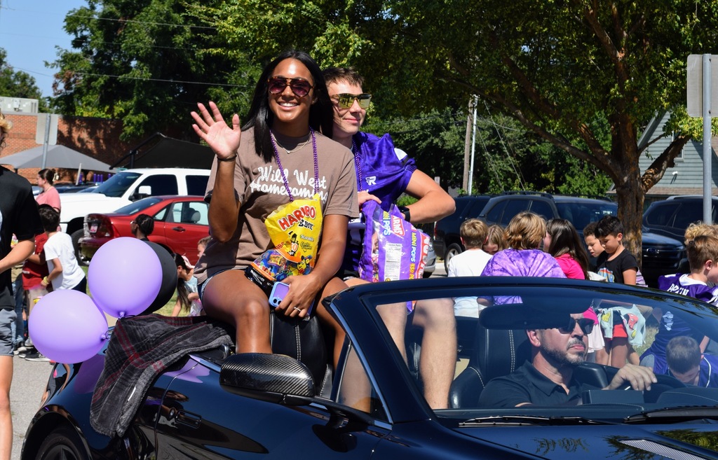 A young woman in sunglasses and a brown "Wild West Homecoming" t-shirt sits on the back of a convertible, waving and smiling while holding a bag of Haribo candy. A young man in a purple jersey sits beside her with more candy. A man drives the car, and purple balloons are tied to the back.