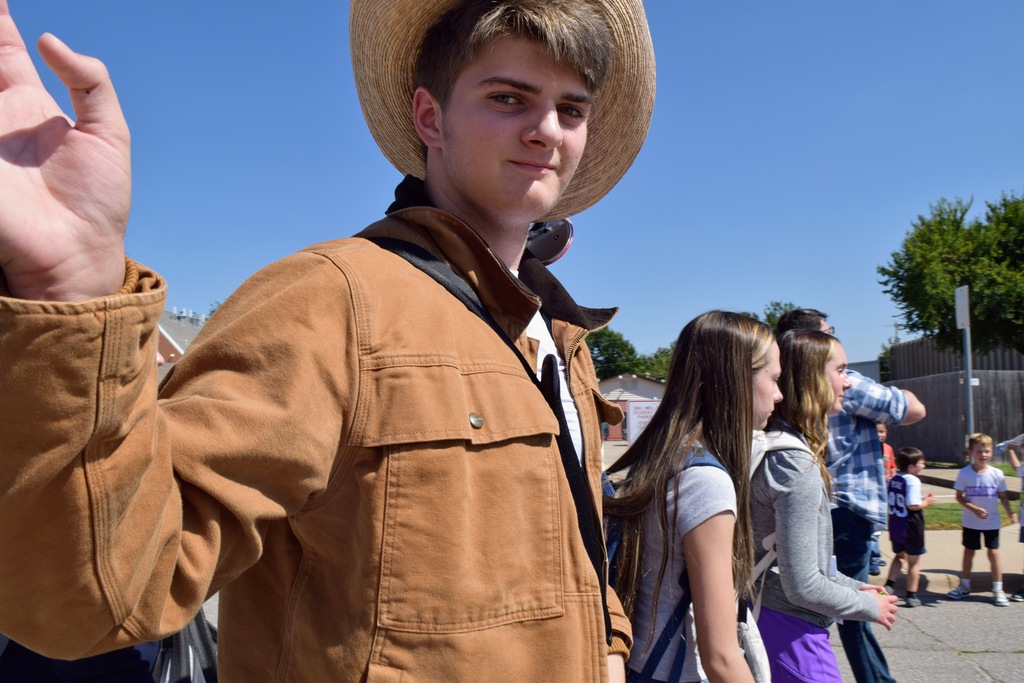 A young man in a tan jacket and straw cowboy hat walks in the parade, raising his hand in a wave. Behind him, more students walk along the street, and some children watch from the sidewalk.