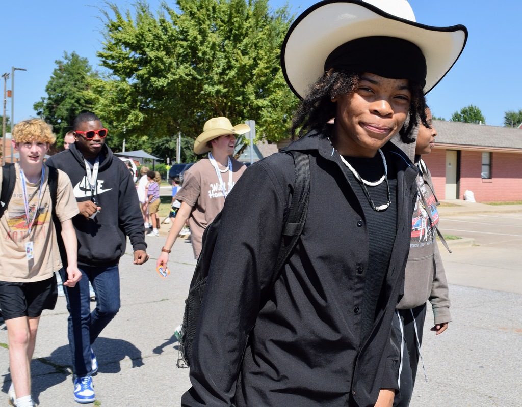 A group of high school students walk together outside during a parade. The student in front wears a large white cowboy hat, a black shirt, layered necklaces, and a backpack, smiling at the camera. Other students walk behind him, including one in a black Nike hoodie with red sunglasses and another in a tan cowboy hat.