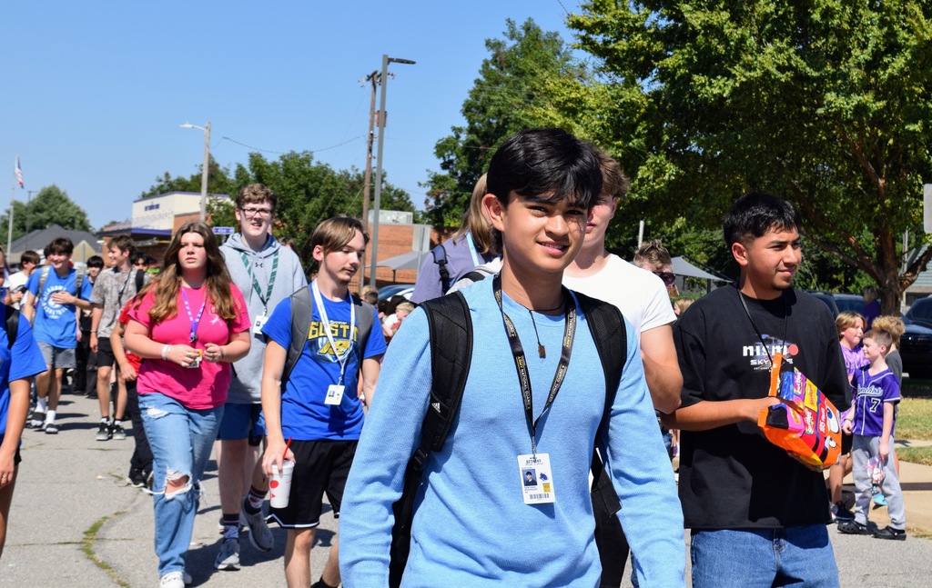 A group of high school students walk in the parade. The student in the foreground smiles in a blue long-sleeve shirt and black backpack. Other students follow behind him, wearing blue and black shirts, carrying drinks and bags of candy.