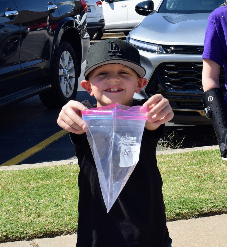 A young boy wearing a black LA Dodgers hat holds up an empty plastic Ziploc bag with a big grin, standing on the grass near parked cars.
