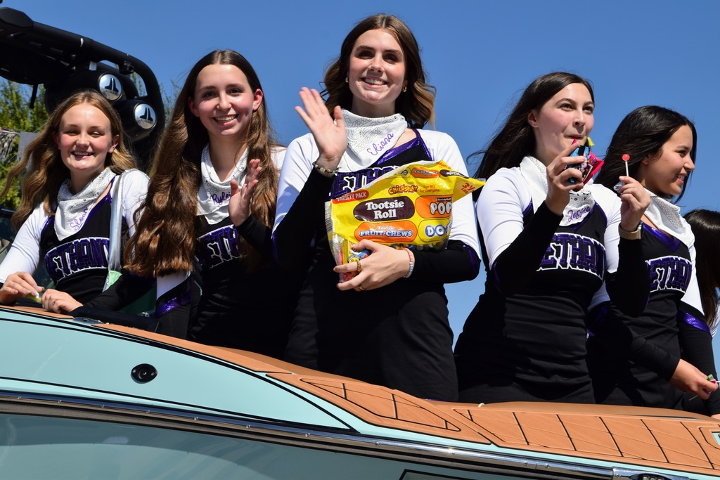 Five cheerleaders in matching black, purple, and white uniforms ride on a parade float. They smile and wave at the crowd while holding bags of candy. One girl waves while another holds up a lollipop.