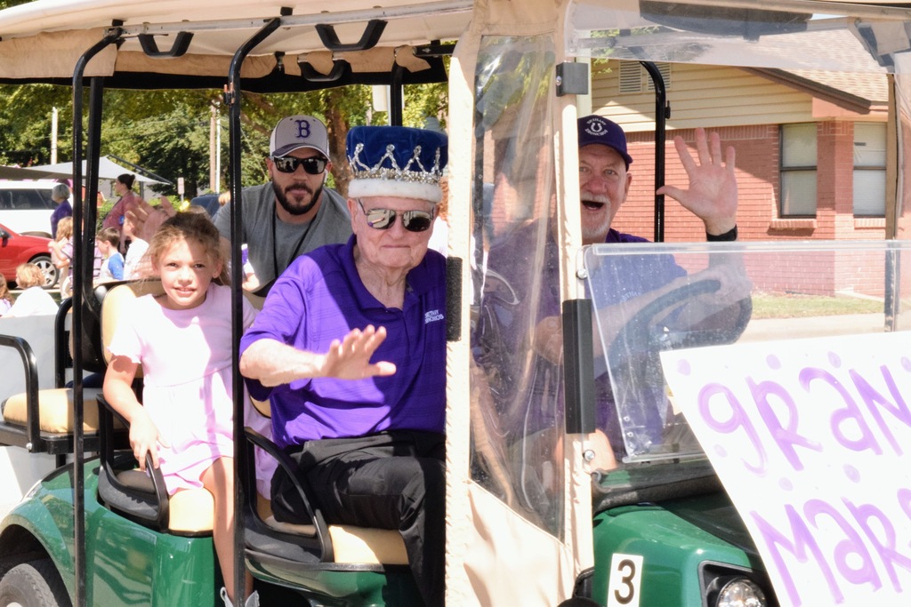 An older man wearing a crown and sunglasses rides in a golf cart labeled “Grand Marshal.” He waves to the crowd while sitting next to a young girl in a pink dress. Two men ride with him, one in a Bethany hat and the other in sunglasses.