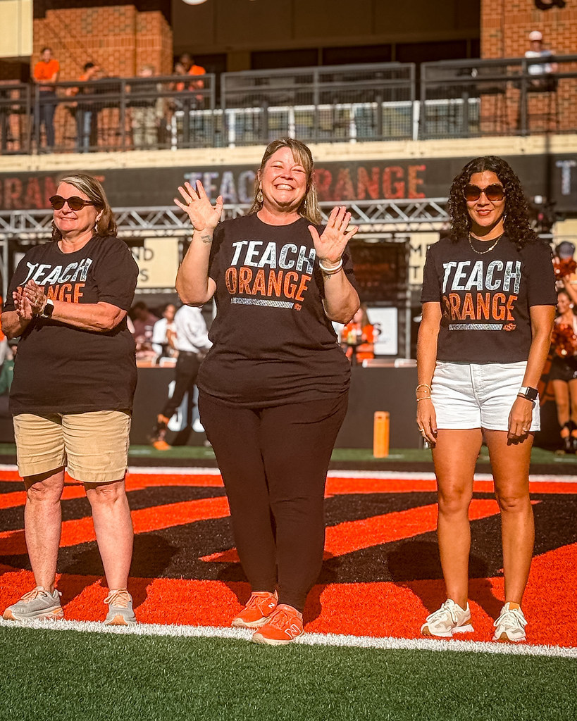 Miss McCann stands on the football field at OSU between two other teachers, smiling and waving as they are recognized during the game.