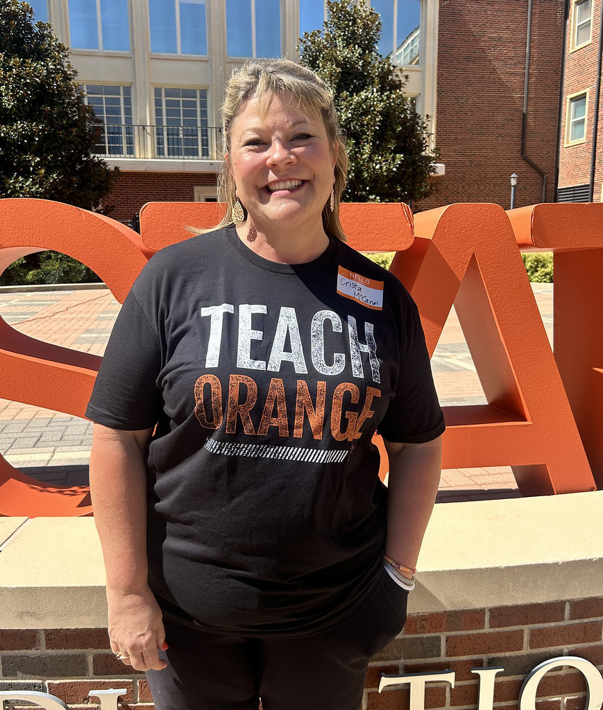 Miss McCann smiles in front of large orange OSU letters on campus, wearing a black “Teach Orange” shirt.