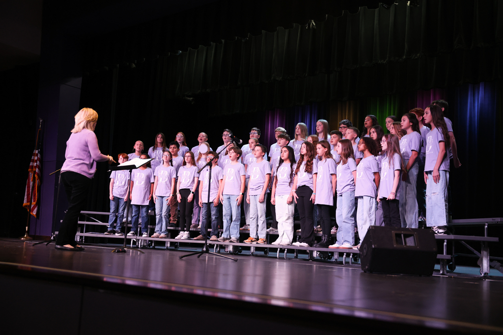photo of the sixth grade choir standing on risers on stage performing. Students wearing lavendar t-shirts, director in front