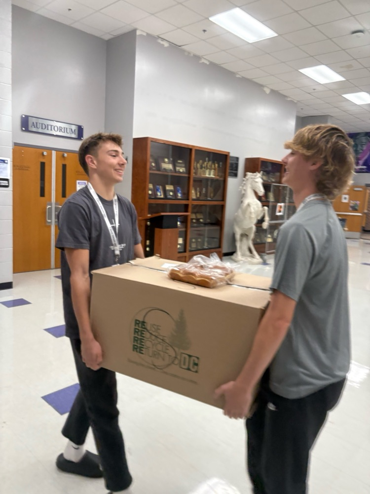 Two high school students walk down a school hallway smiling as they carry a large cardboard box together. The hallway has trophy cases, an auditorium entrance, and a white horse statue in the background.