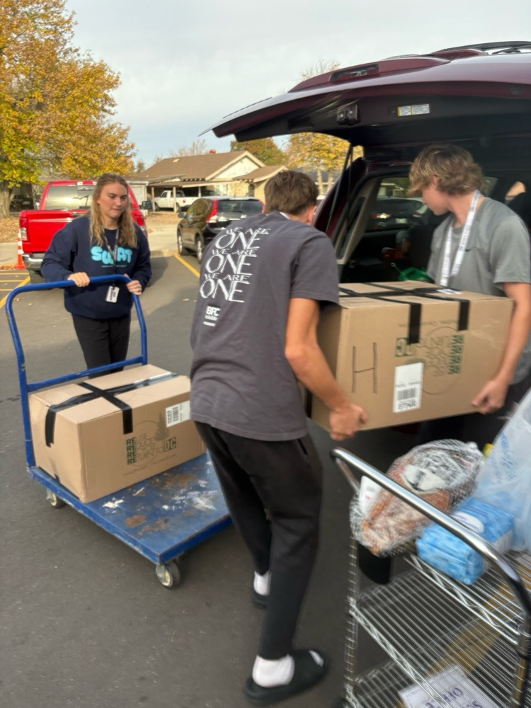 Three students load large cardboard boxes into the back of a maroon SUV. One student pushes a blue cart with more boxes, and another metal cart holds groceries including eggs and packaged meat.