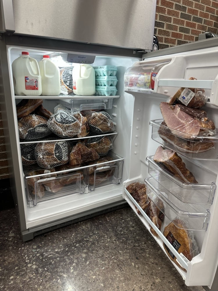 A refrigerator packed full of donated food, including multiple hams in netting, several cartons of eggs, and gallons of milk. The shelves and door compartments are completely filled.