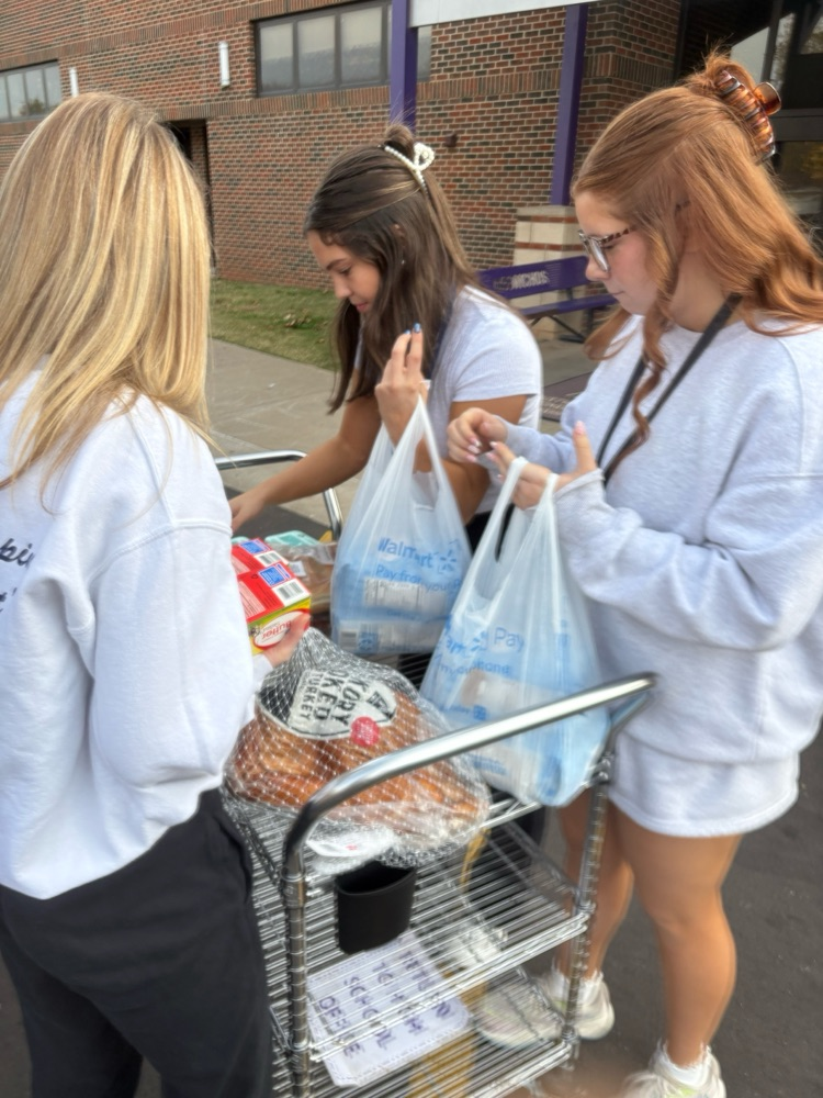 Three high school students sort groceries on a metal cart outside. They hold plastic bags filled with food, and packaged hams sit on the lower shelf. A brick school building is in the background.